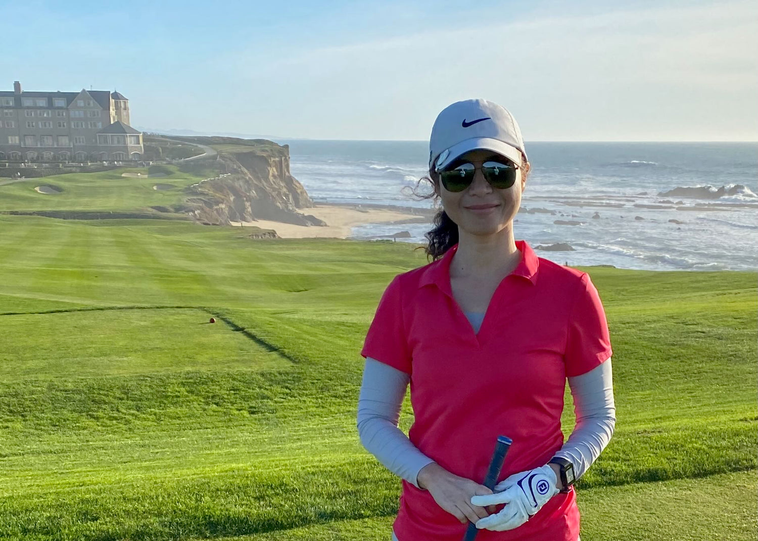 Woman in a red shirt and sunglasses holding a golf club on a golf course with ocean view
